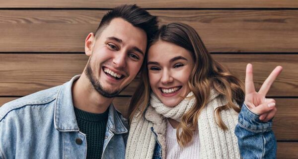 cheerful-european-girl-knitted-scarf-posing-with-peace-sign-wooden-wall-indoor-shot-laughing-married-couple-having-fun-together