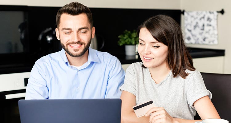 miling young couple sitting kitchen table using laptop while paying bills